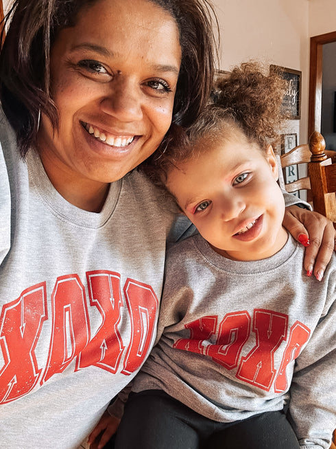 Mom and daughter wearing matching Valentine’s Day sweatshirts that say xoxo with red text and heather grey sweatshirts
