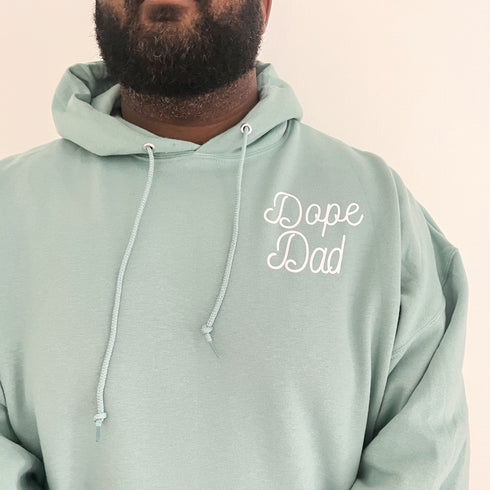 black dad behind a white background modeling an ocean blue color hoodie with screen print design that says dope dad, cute mens sweatshirt hoodie